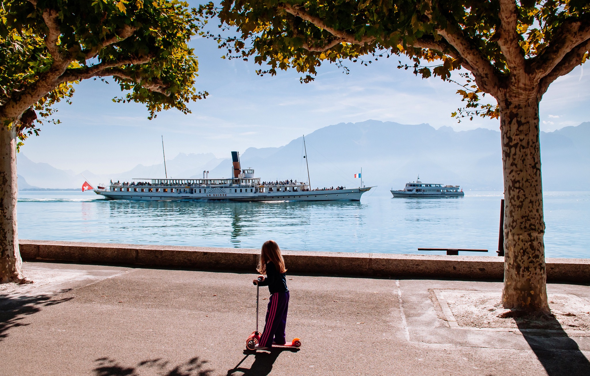 SEP 25, 2013 Montreux, Switzerland - Vintage sightseeing boats  in lake Geneva and kid riding kick scooter on bright blue sky day in autumn with Swiss alps in background